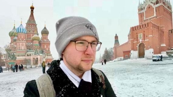 A photo of a British man, wearing glasses and a hat, standing in Russia with an ornate church behind him